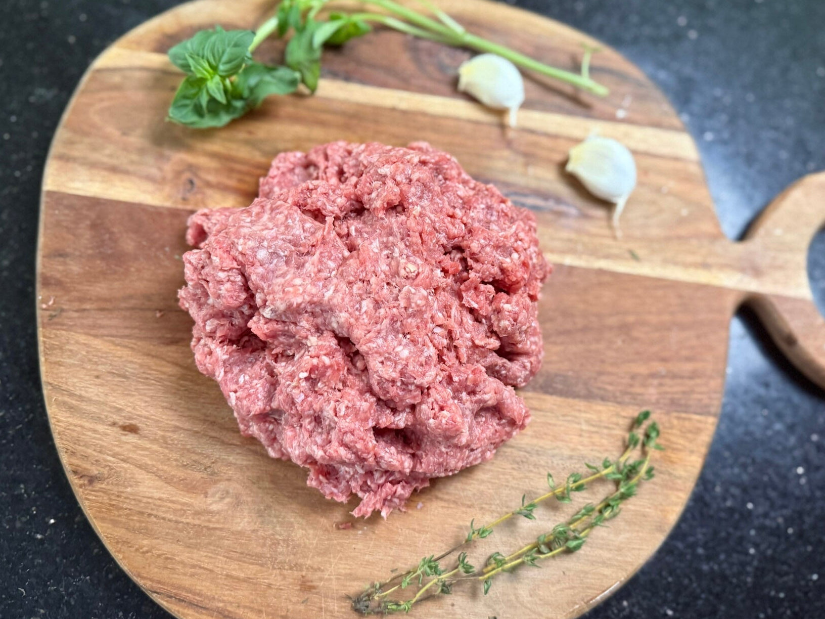 Grass Fed ground meat on a wooden cutting board with herbs on a black surface