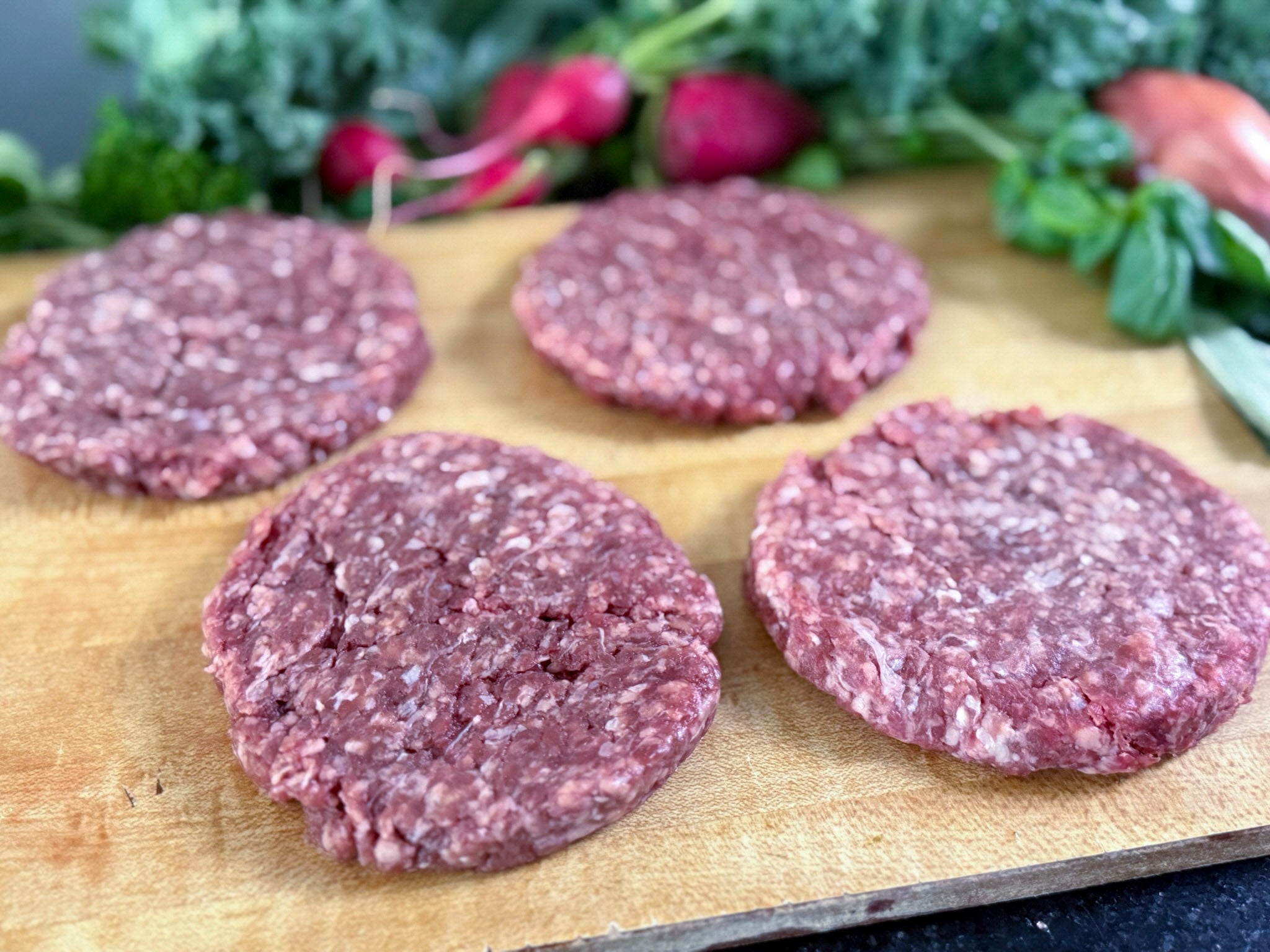 Four grass fed raw hamburger patties on a wooden cutting board with vegetables in the background.