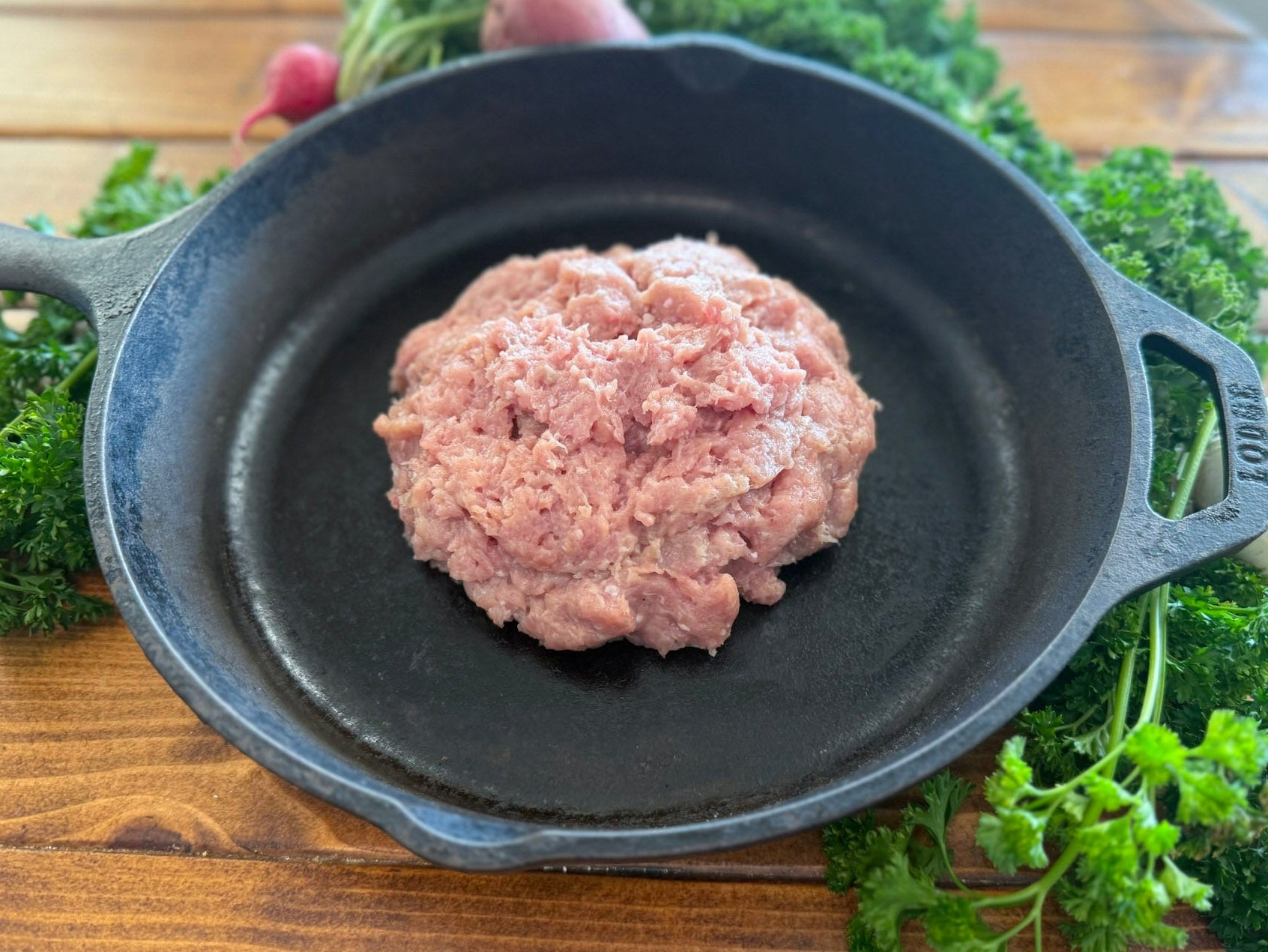 Uncooked pasture raised Italian spice ground chicken in a cast iron skillet on a wooden surface with herbs.