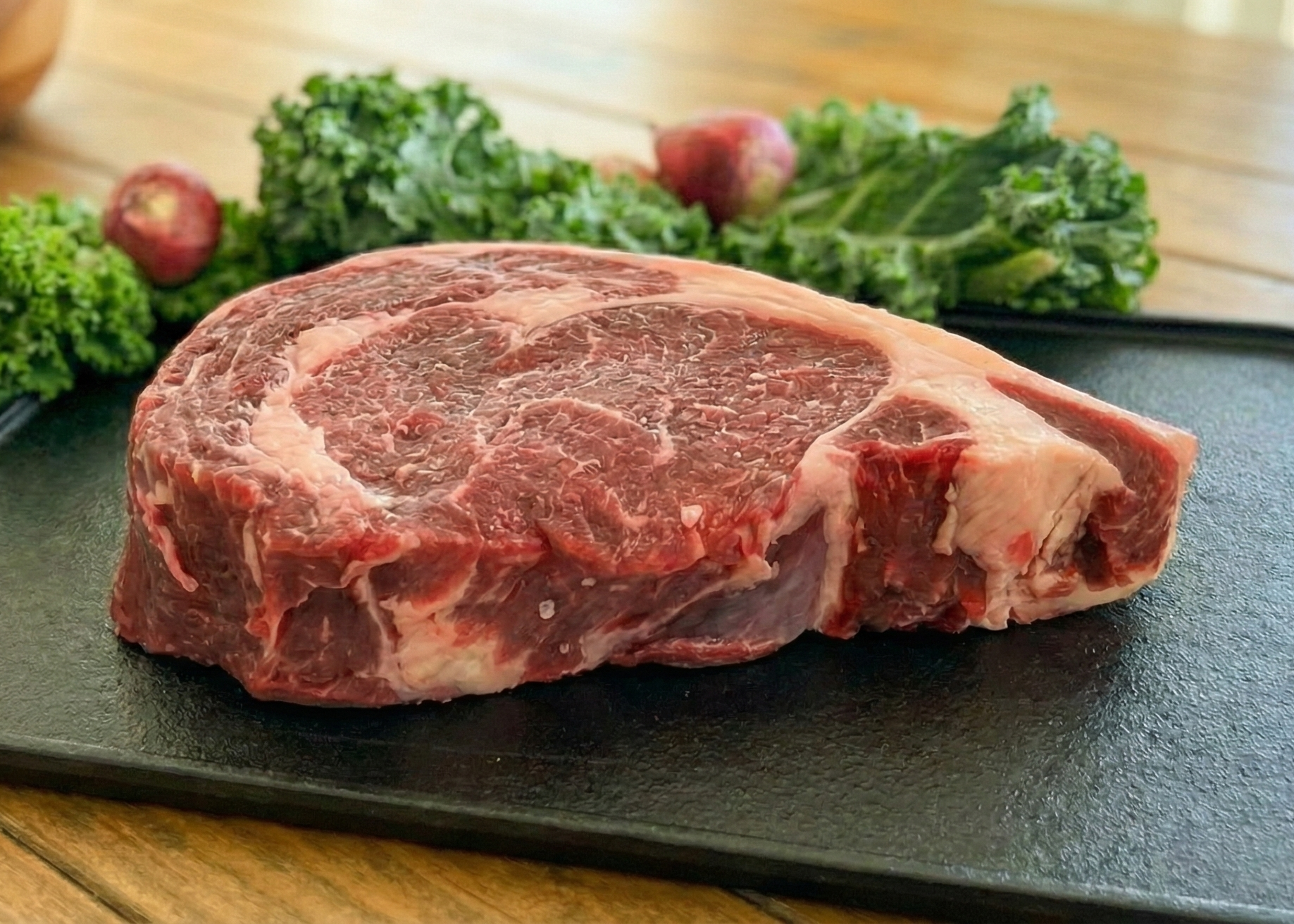 Beef Single New York Strip Steak (Single) on a black cutting board with greens on a wooden surface