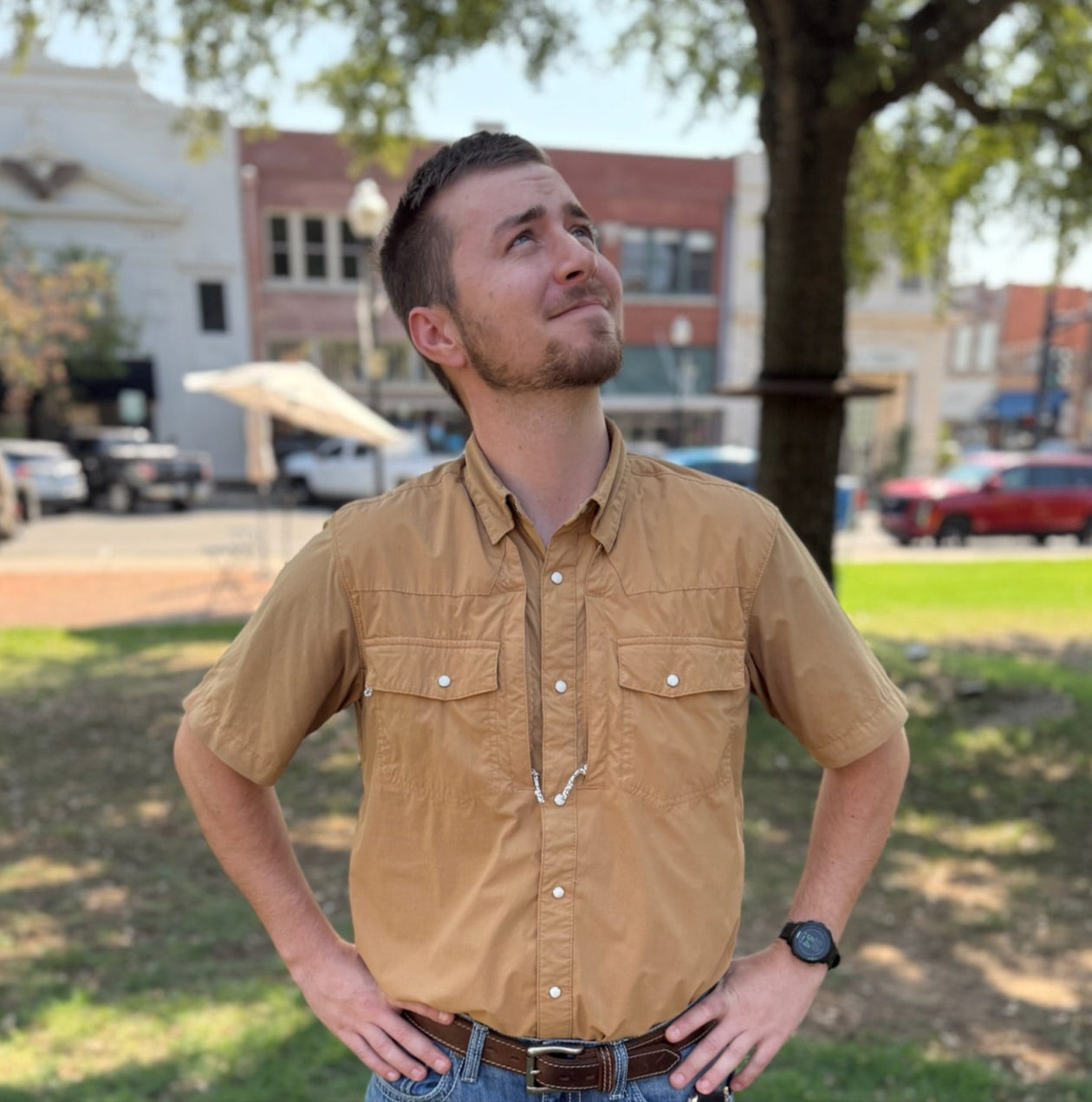 Man wearing a brown shirt and blue jeans standing in a park with buildings in the background