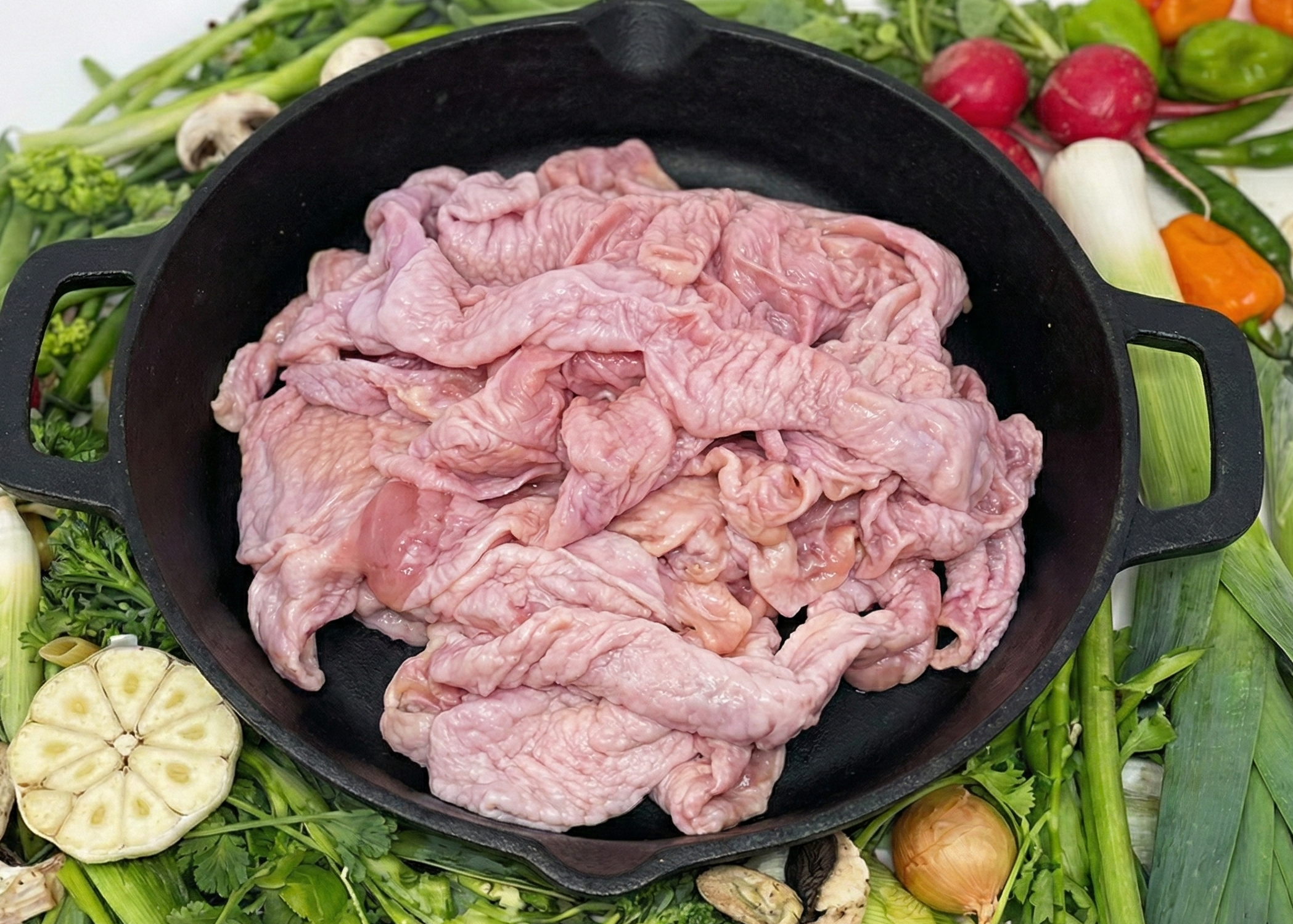 Raw Chicken Skin with Fat in a black skillet surrounded by green vegetables on a white background