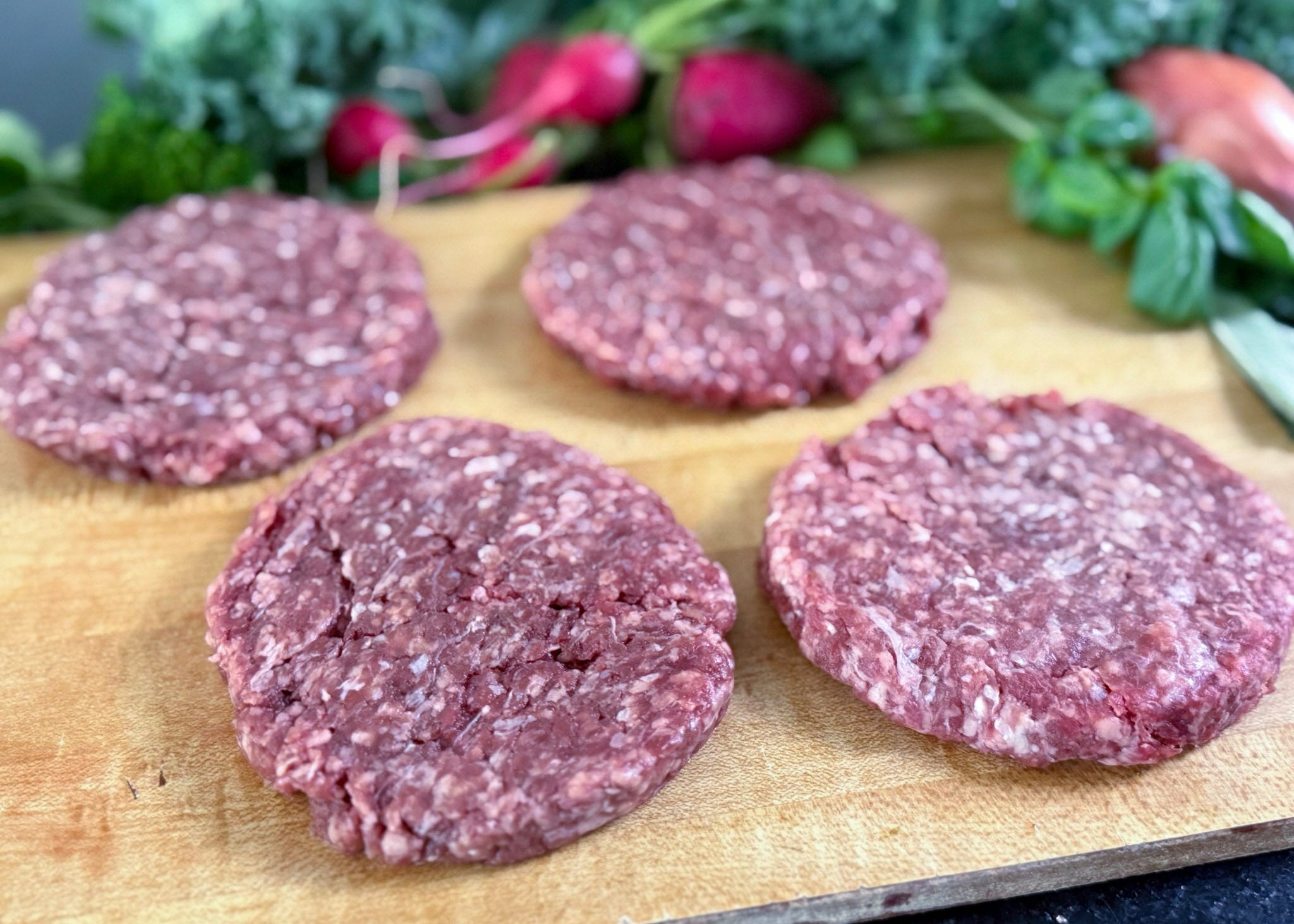 Ground Beef High Fat Quarter-Pound Patties on a wooden cutting board with vegetables in the background.