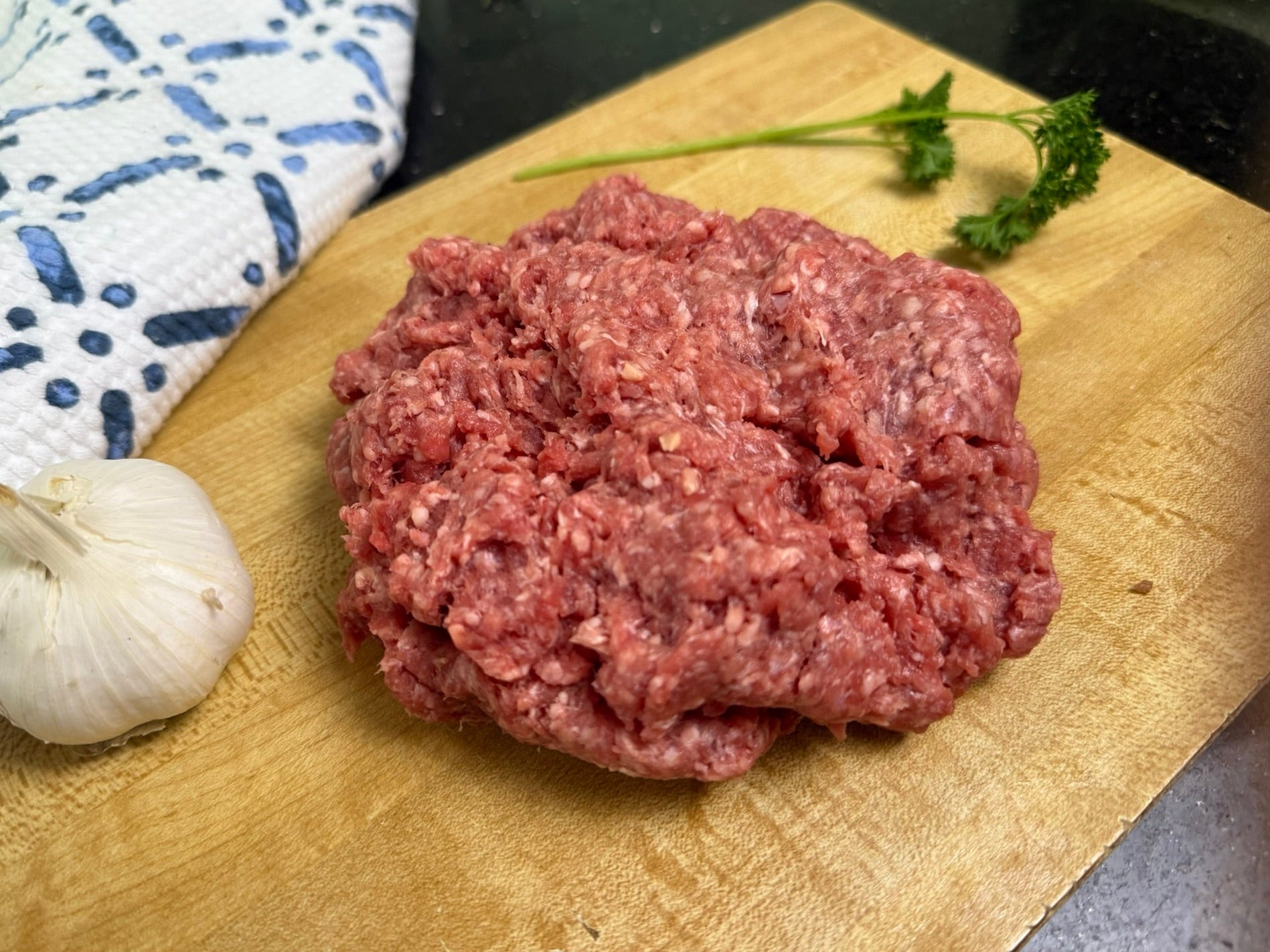 Raw ground meat on a wooden cutting board with a bulb of garlic and parsley.