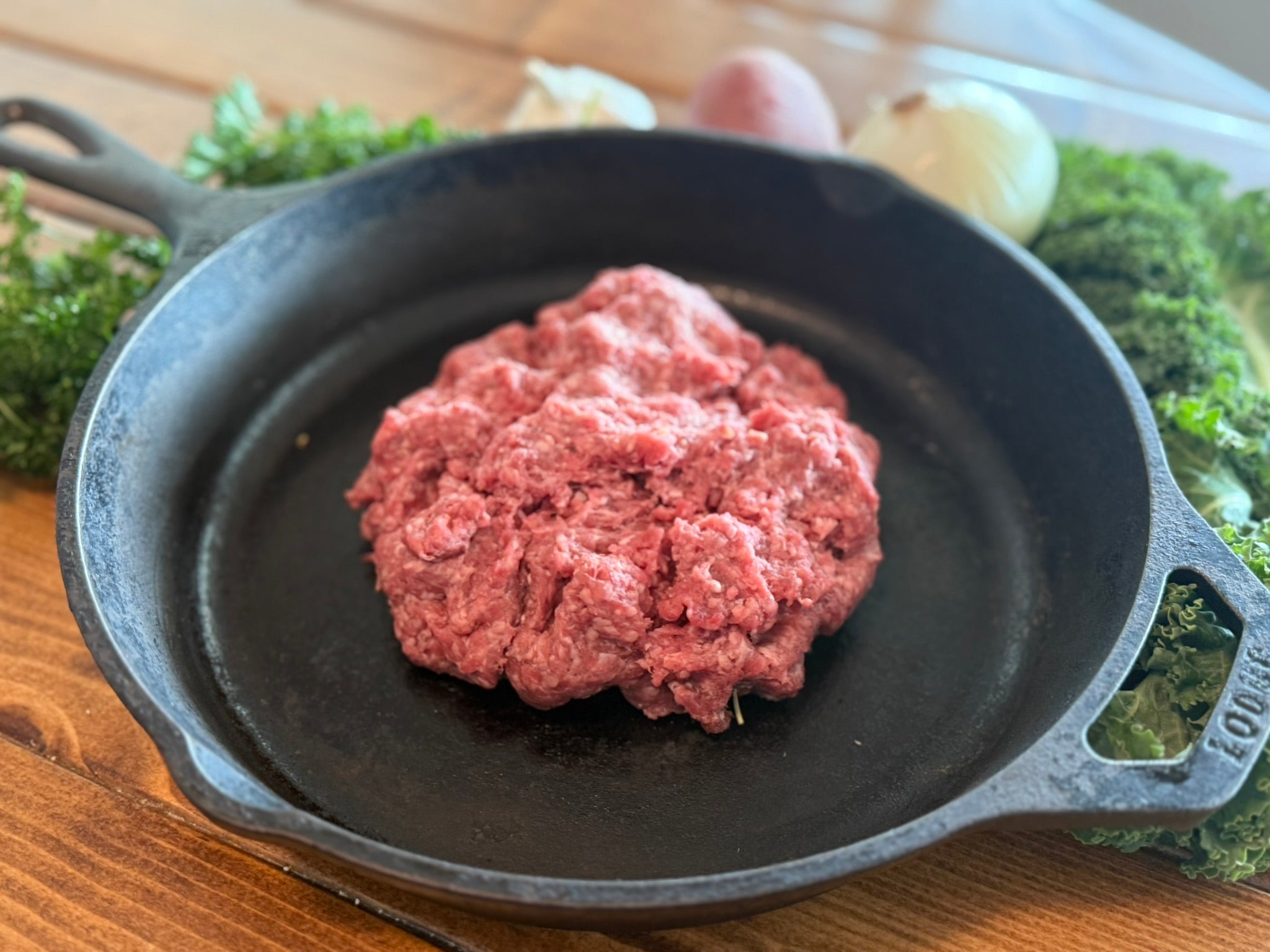 Raw ground meat in a cast iron skillet on a wooden surface with greenery in the background
