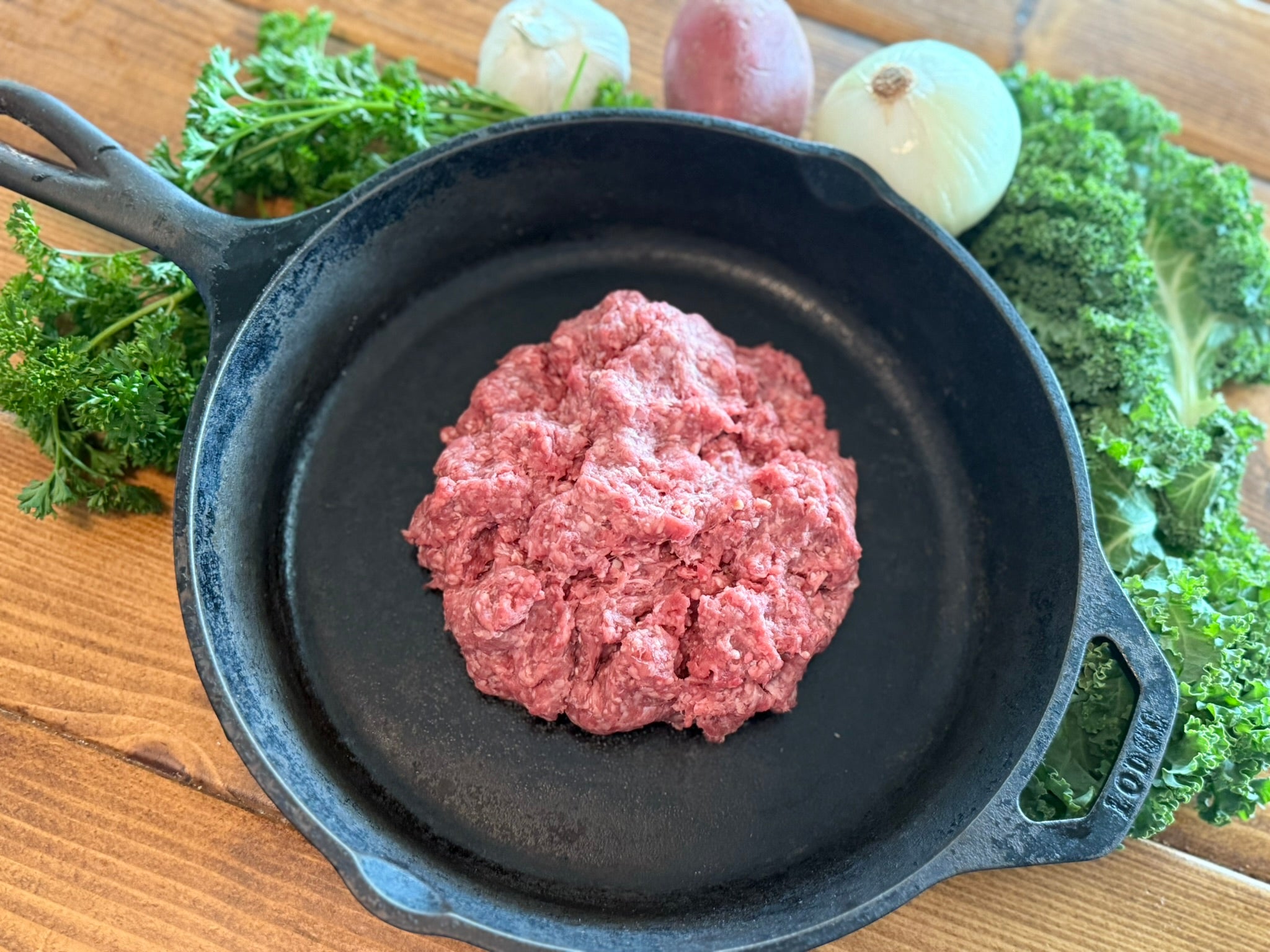 Raw ground meat in a cast iron skillet with herbs and onions on a wooden surface