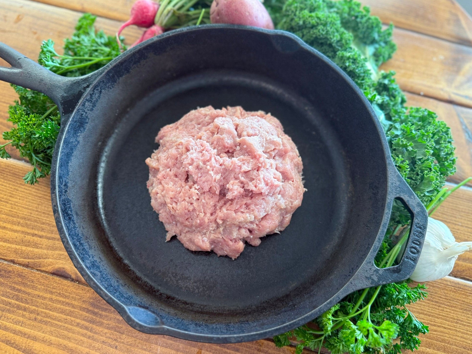 Raw ground pork  in a cast iron skillet on a wooden surface with vegetables around