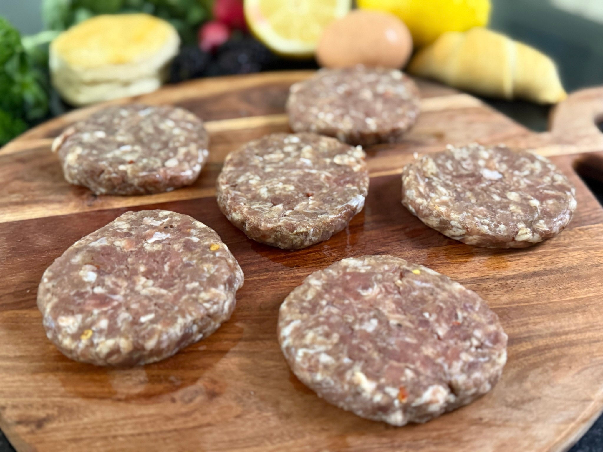 Raw ground sausage patties on a wooden cutting board with lemons and vegetables in the background.