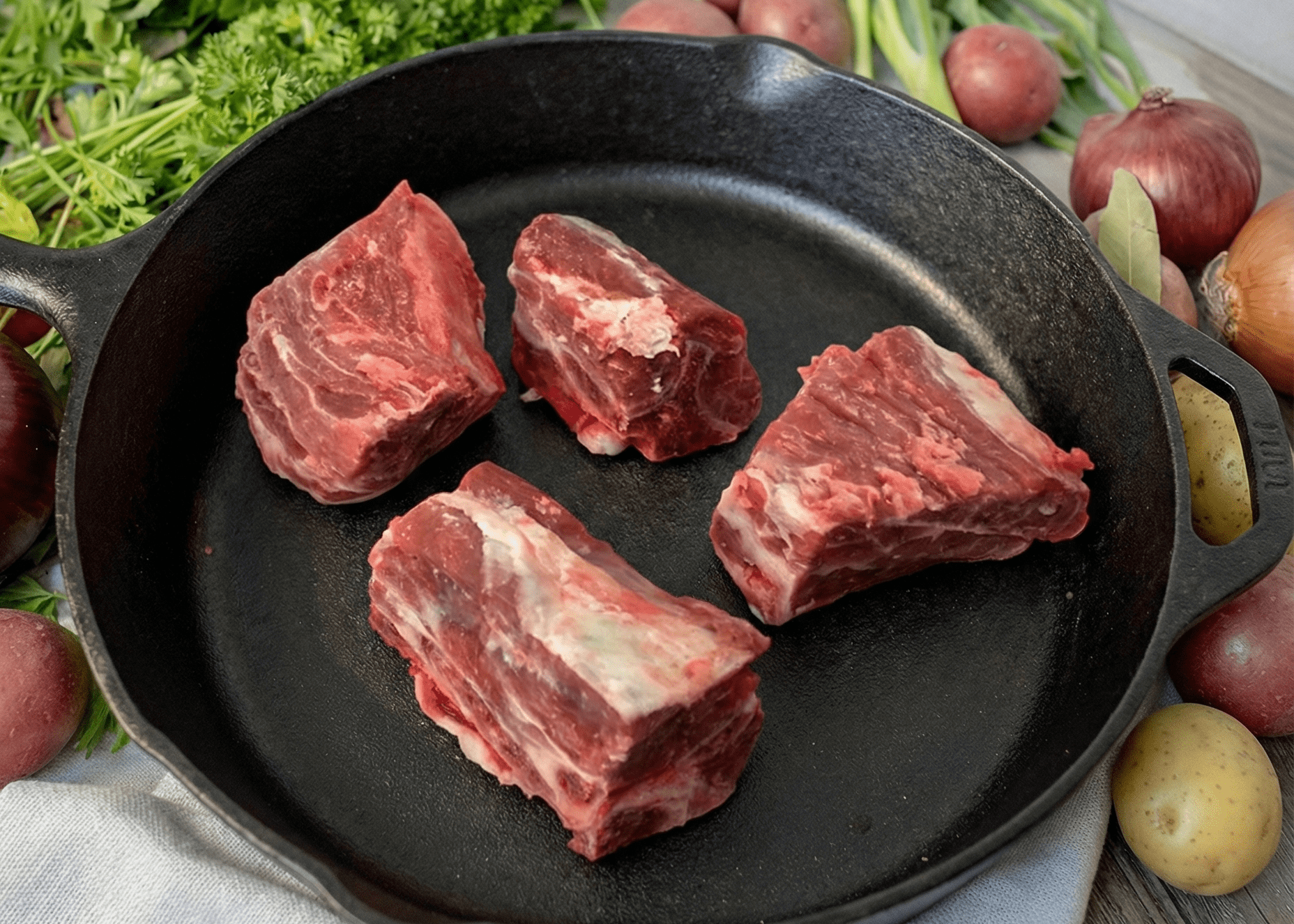 Raw Lamb Bone-In Stew Meat in a black cast iron skillet with vegetables on a wooden surface