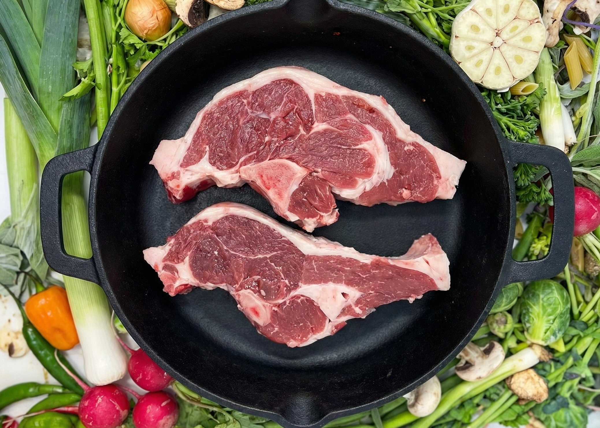 Two raw Lamb Sirloin Chops in a cast iron skillet surrounded by fresh vegetables on a white background