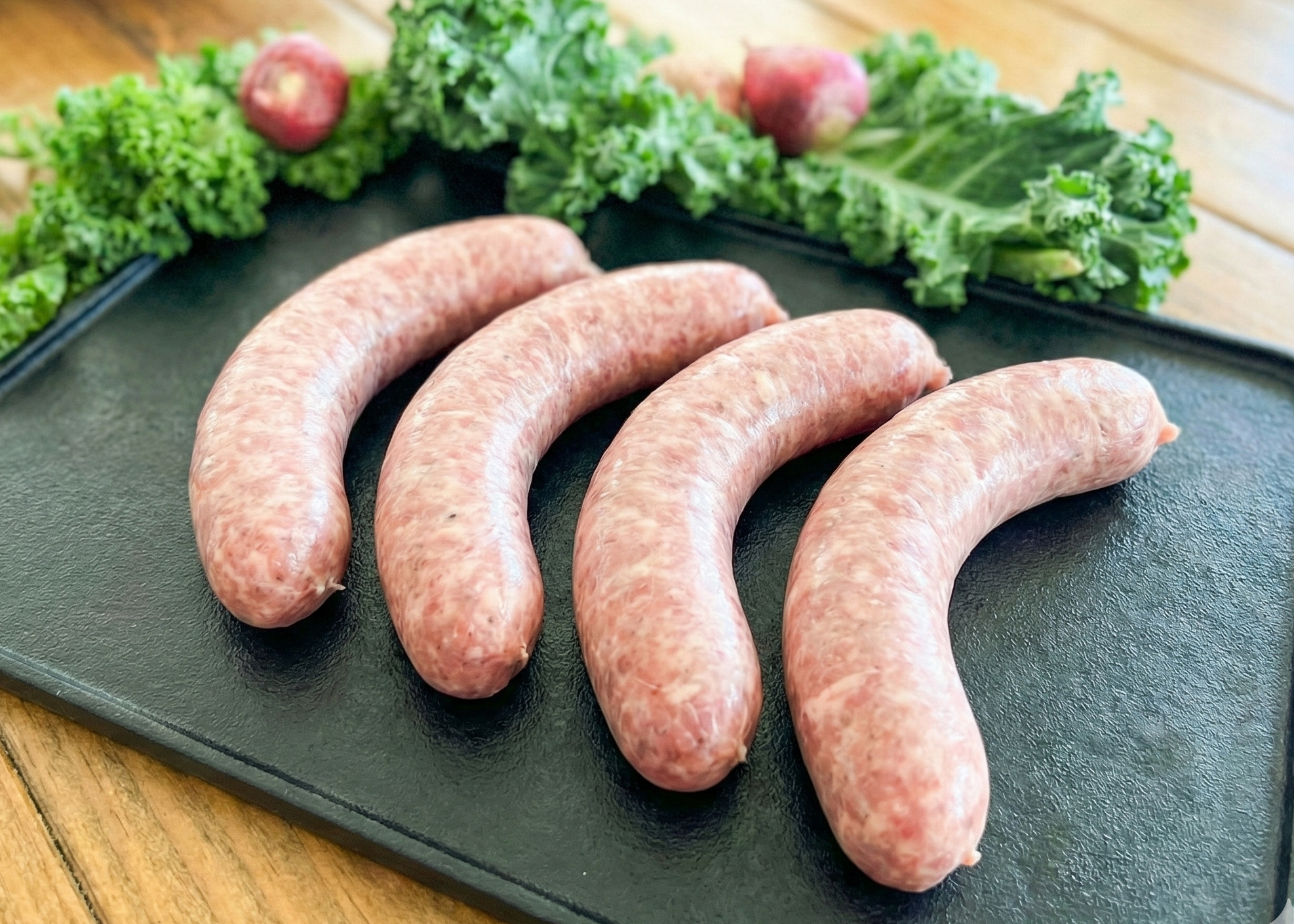 Raw Natural Pork Bratwurst on a black slate surface with green leafy vegetables on a wooden table.