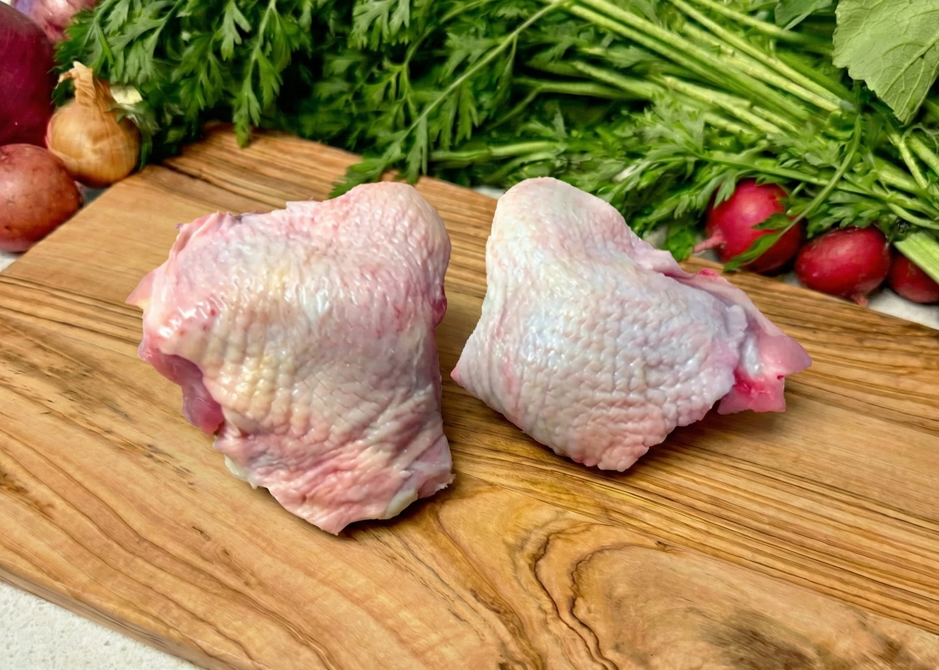 Two raw Omega-3 Chicken Bone-In Skin-On Thighs on a wooden cutting board with vegetables in the background.