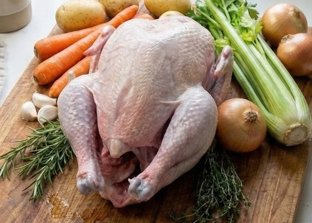 Raw Omega-3 Whole Chicken Roaster on a wooden cutting board with vegetables on a kitchen counter