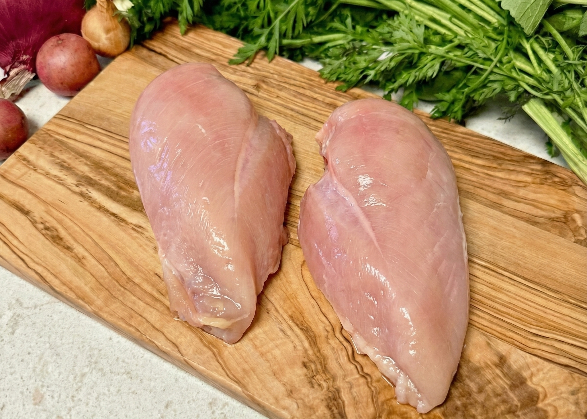 Two raw chicken breasts on a wooden cutting board with vegetables in the background.