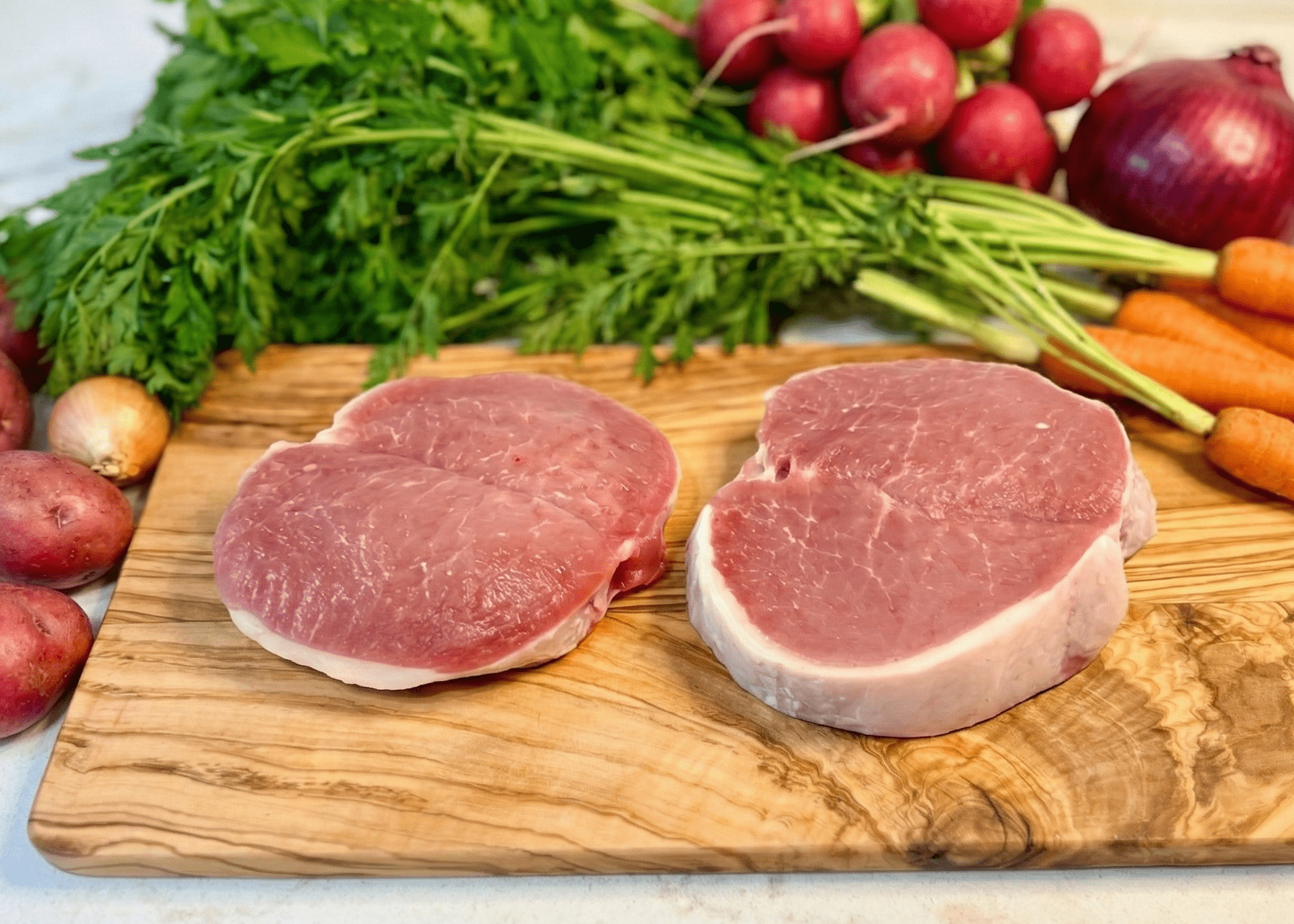 Two raw Pork Butterfly Chops on a wooden cutting board with vegetables in the background.