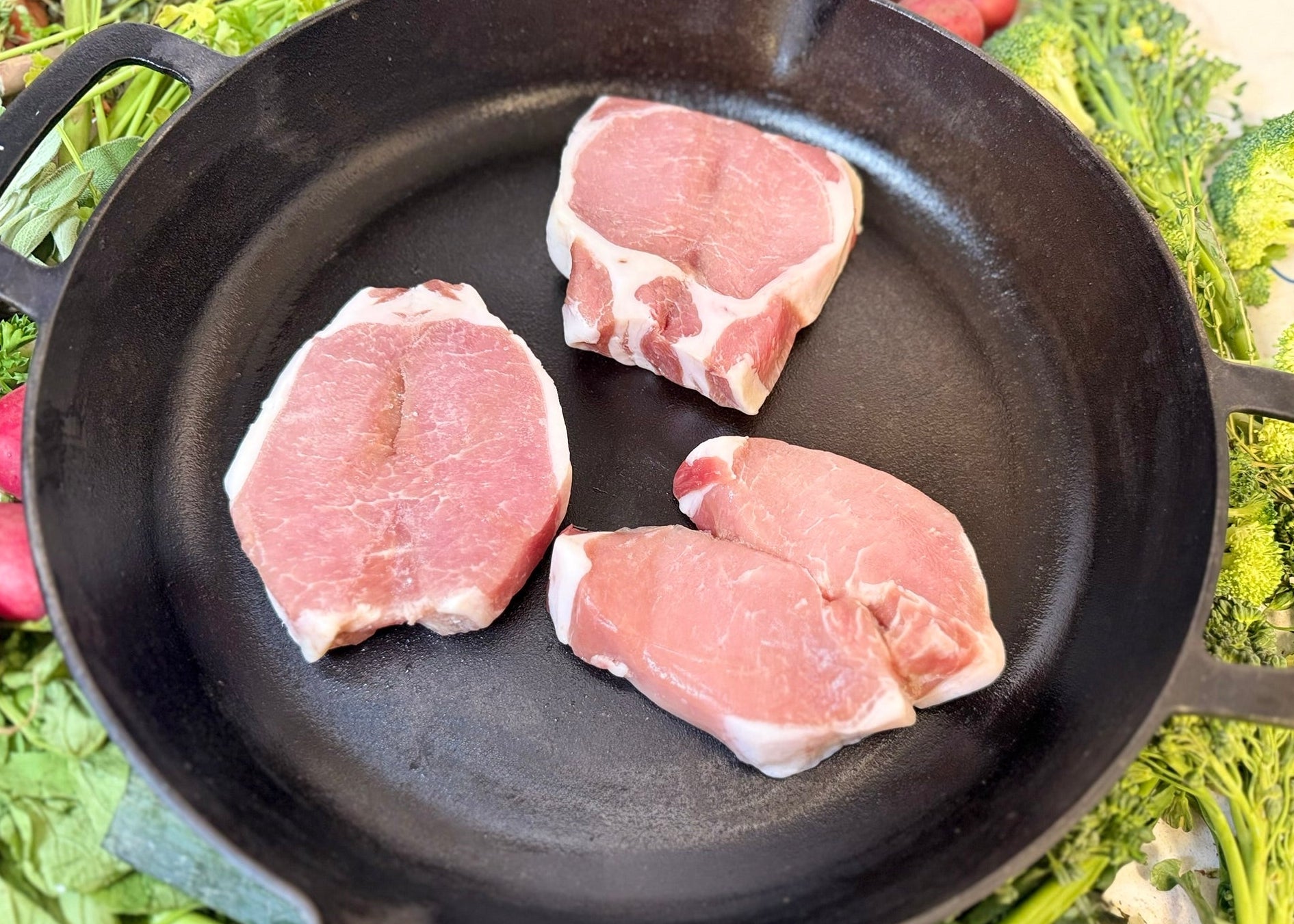 Three raw pork chops in a black cast iron skillet with green vegetables in the background