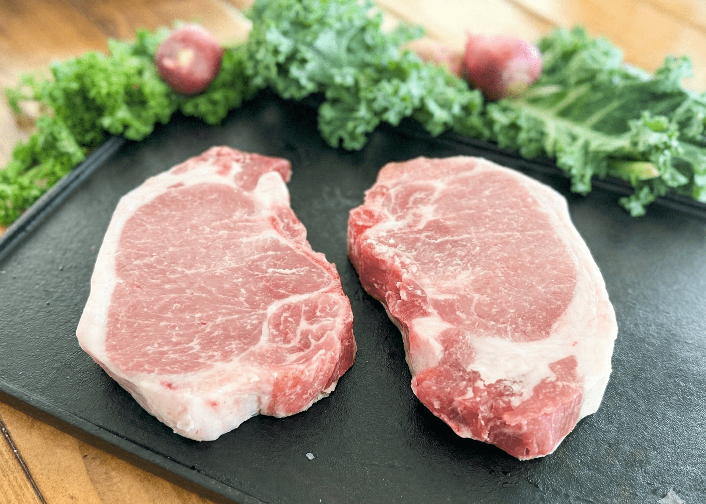 Two raw Pork Loin Chops on a black slate surface with green vegetables in the background.