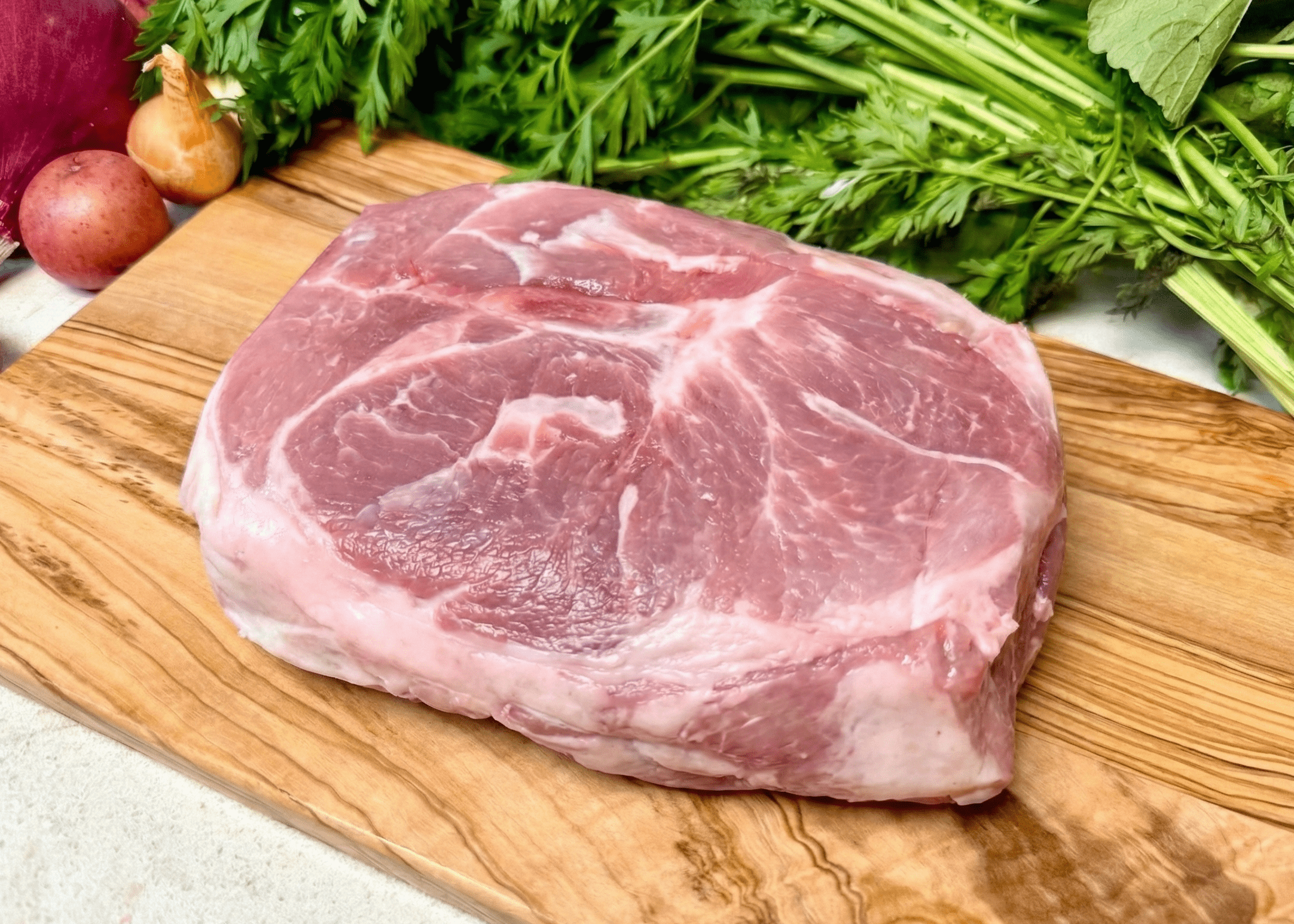 Raw Pork Shoulder (Arm) Roast on a wooden cutting board with vegetables in the background