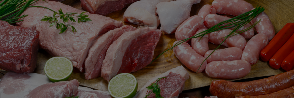 Assorted raw meats including pork and sausages on a wooden board with limes and rosemary.