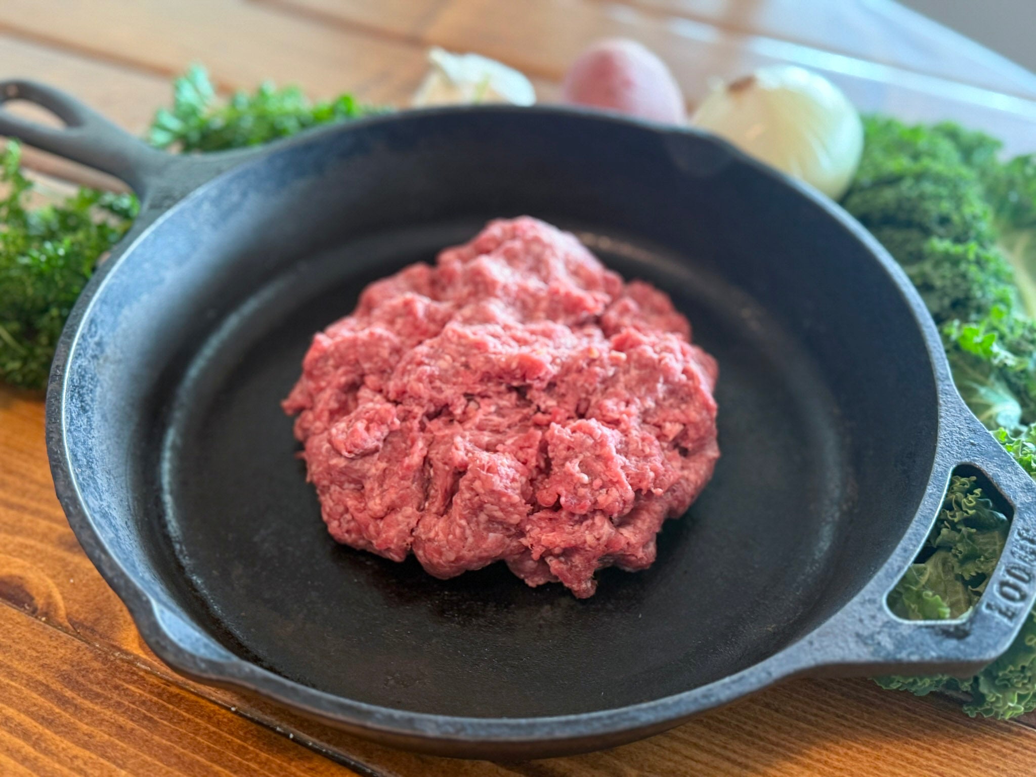 Raw ground meat in a cast iron skillet on a wooden surface with greenery in the background