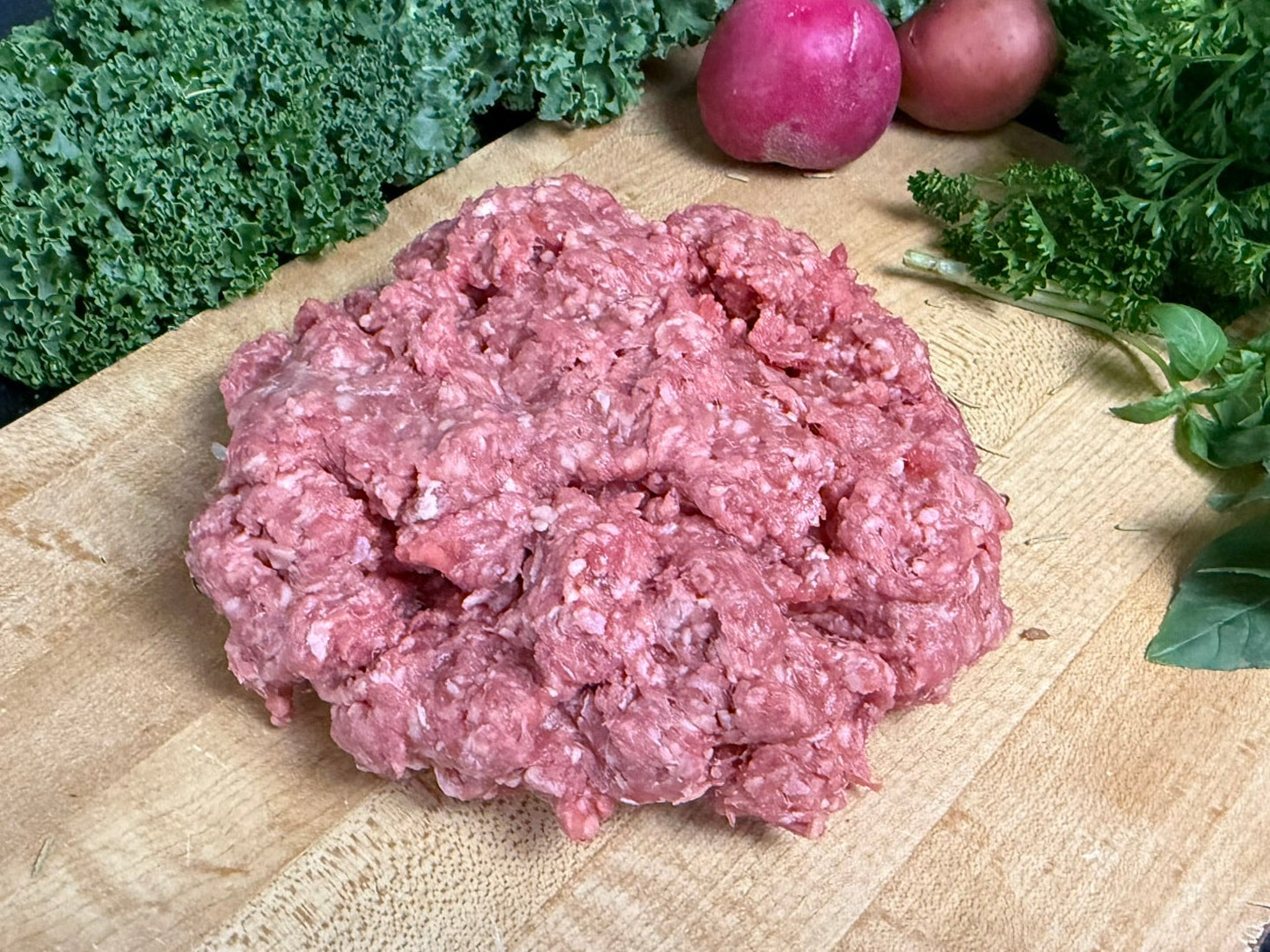 Raw ground meat on a wooden cutting board with kale and radishes in the background.