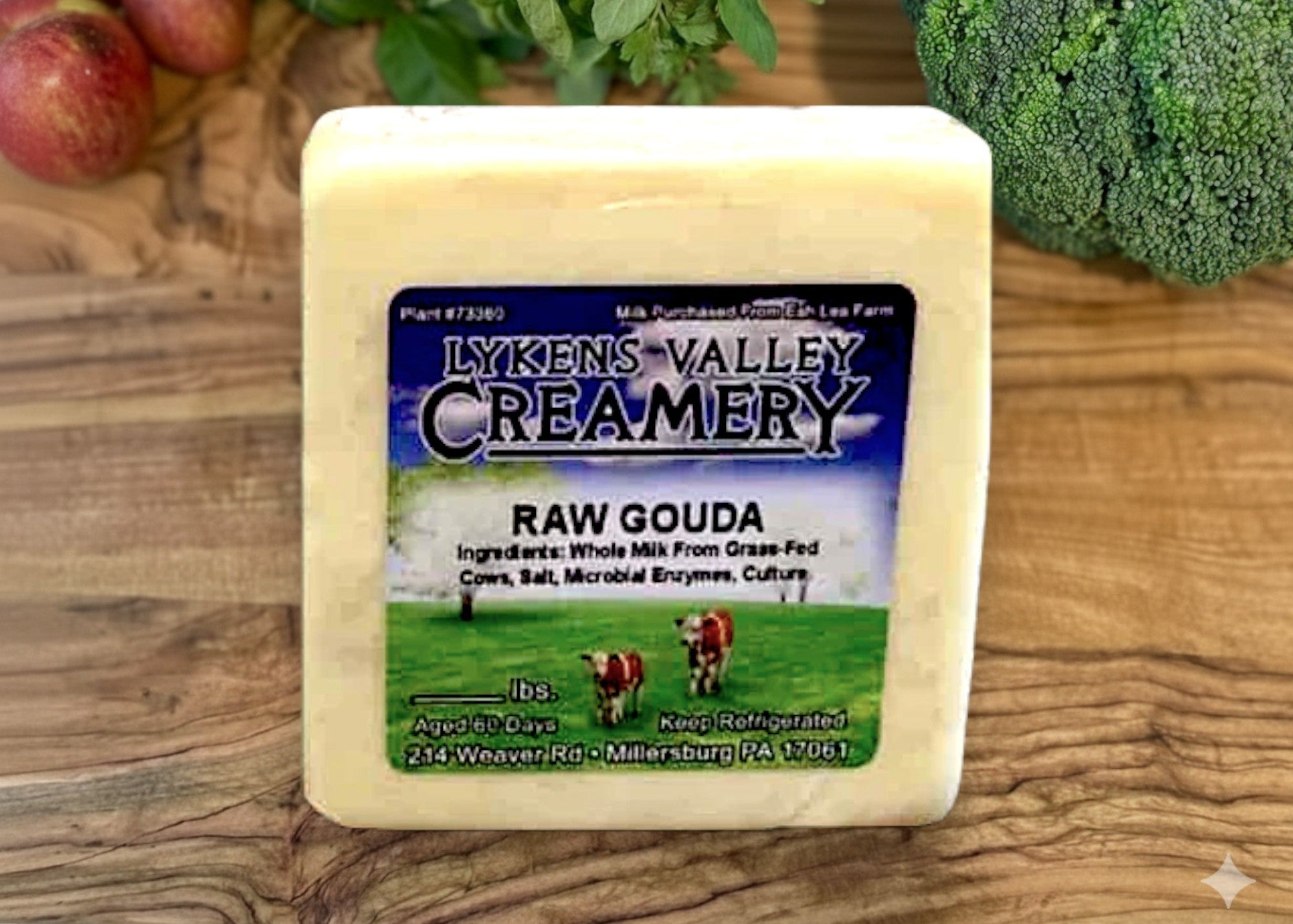 Block of raw gouda cheese from Lykens Valley Creamery on a wooden surface with fruits and vegetables.
