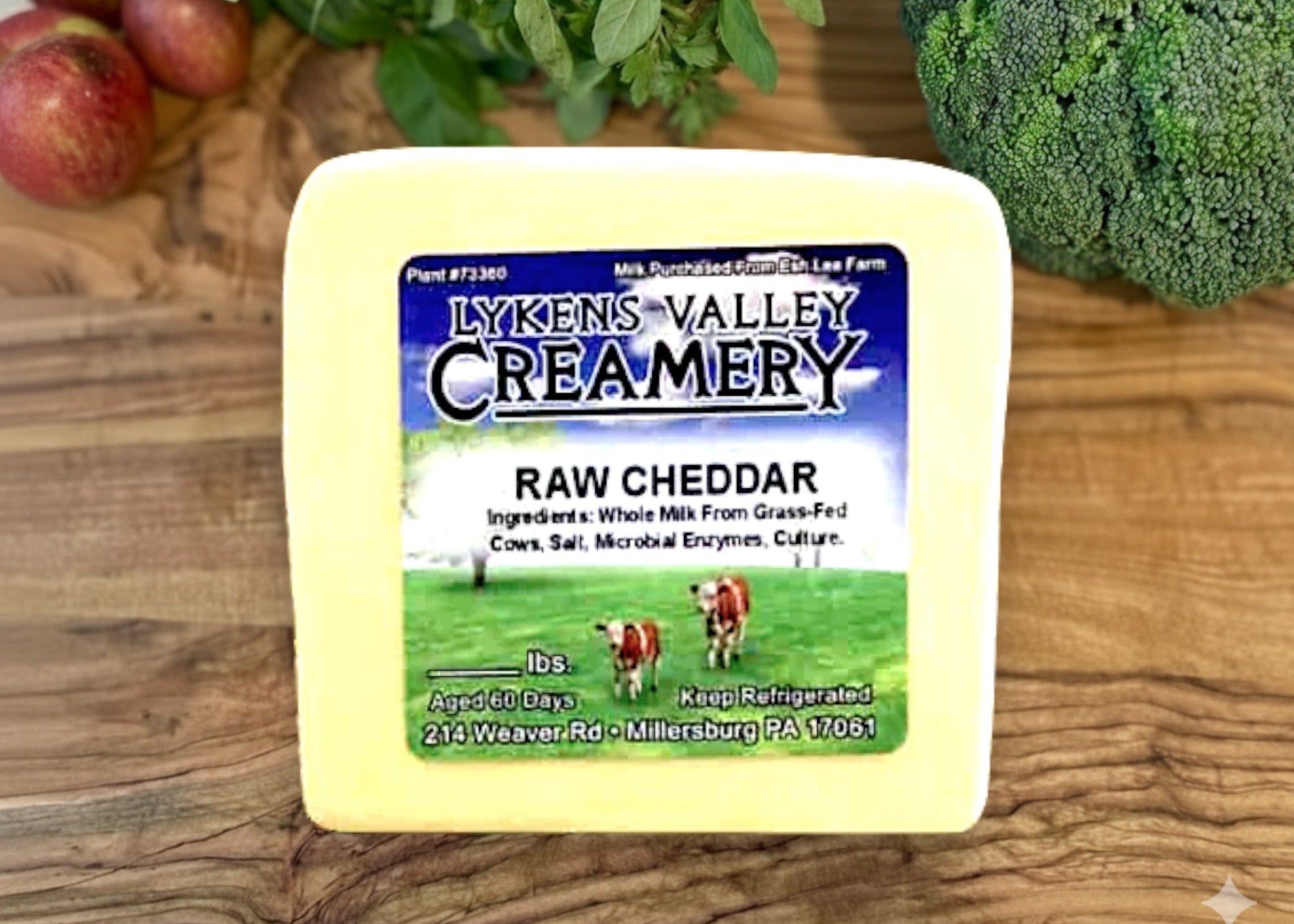 Block of Lyken's Valley Creamery raw cheddar cheese on a wooden surface with fruits and vegetables.