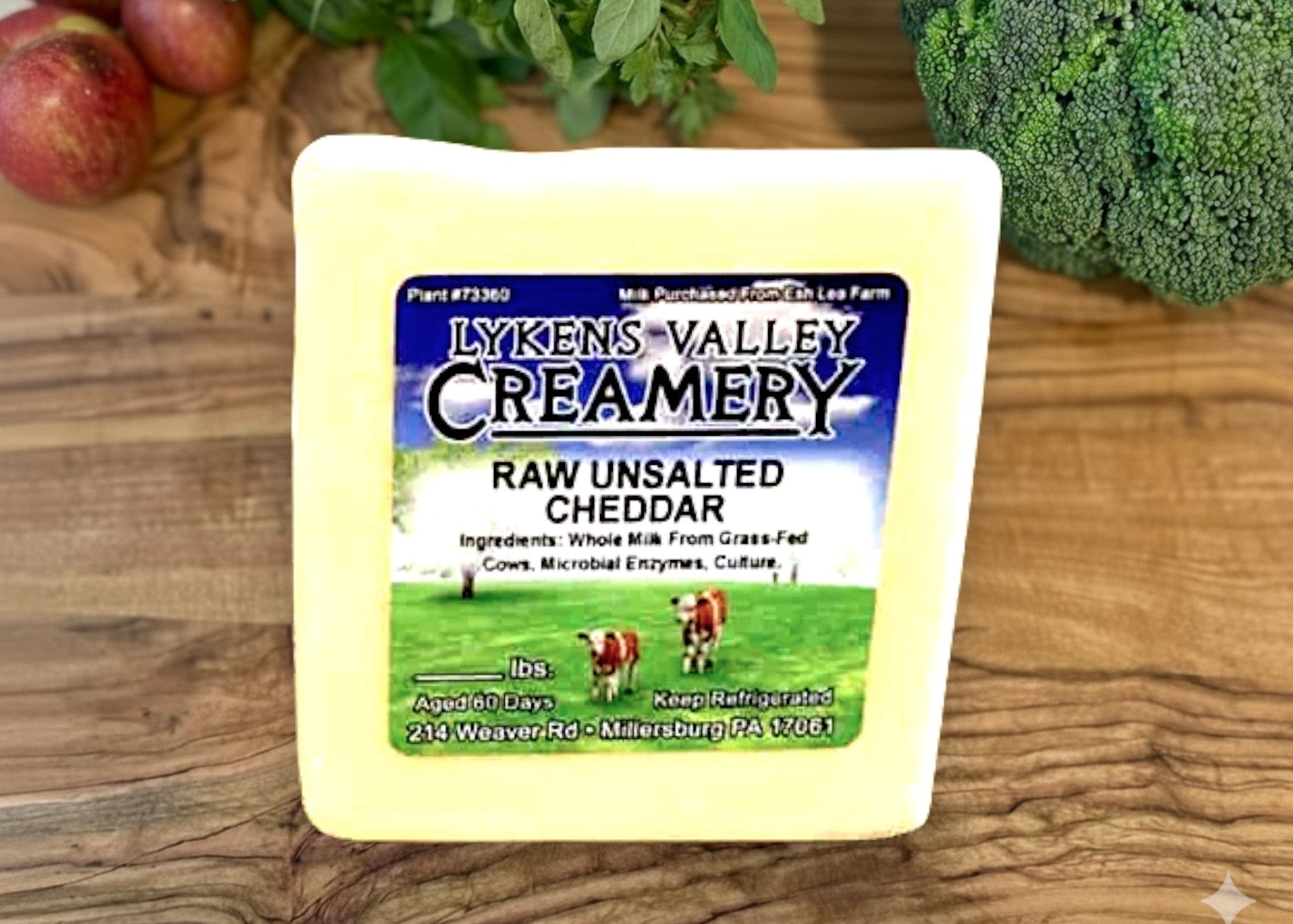 Block of Lyken's Valley Creamery raw unsalted cheddar cheese on a wooden surface with fruits and vegetables.
