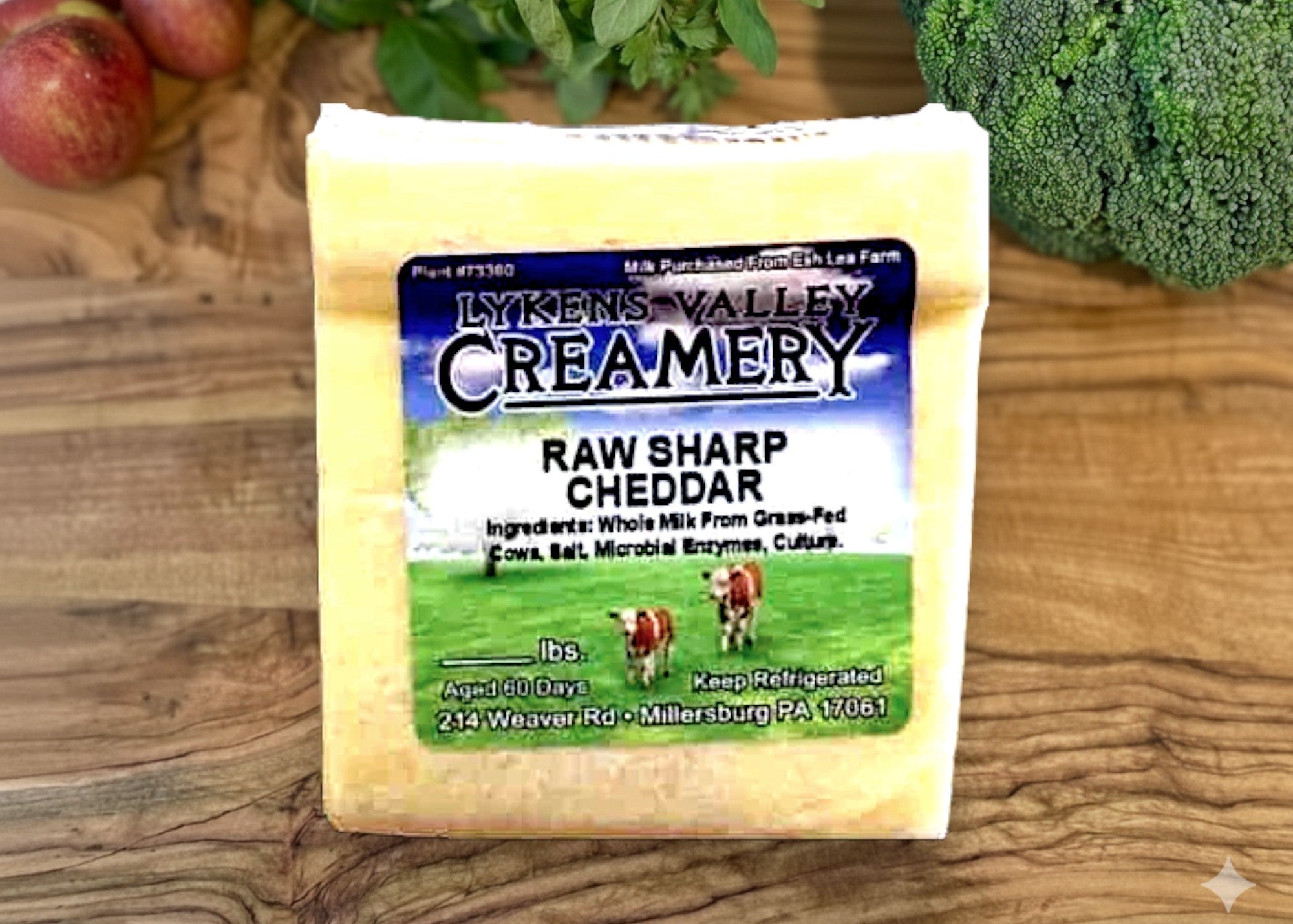 Block of Lyken's Valley Creamery Raw Sharp Cheddar cheese on a wooden surface with vegetables in the background.