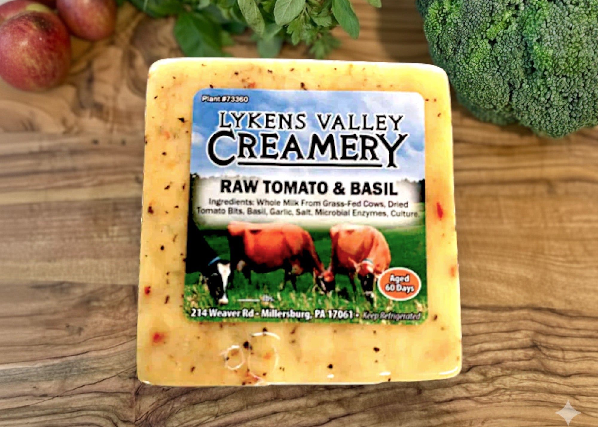 Block of Lyken's Valley Creamery raw tomato & basil cheese on a wooden surface with vegetables in the background.
