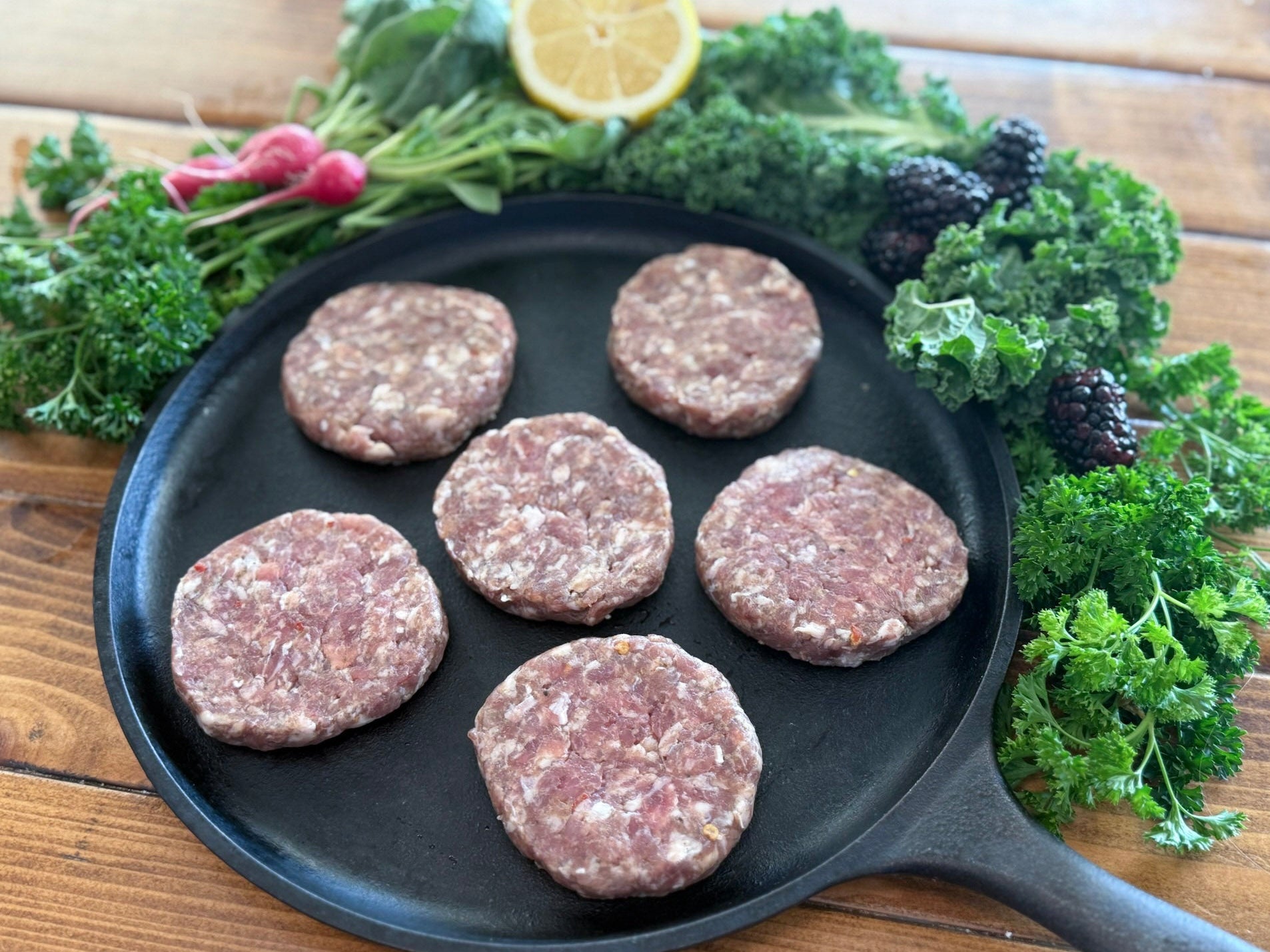 Raw pork sausage patties on a cast iron skillet with fresh greens and a lemon wedge on a wooden surface.
