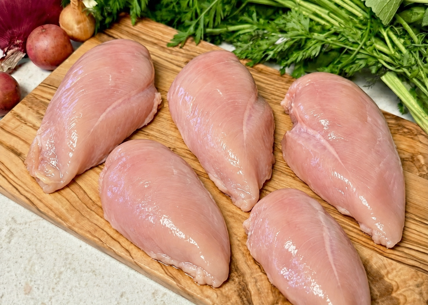 Five raw skinless chicken breasts on a wooden cutting board with vegetables in the background.