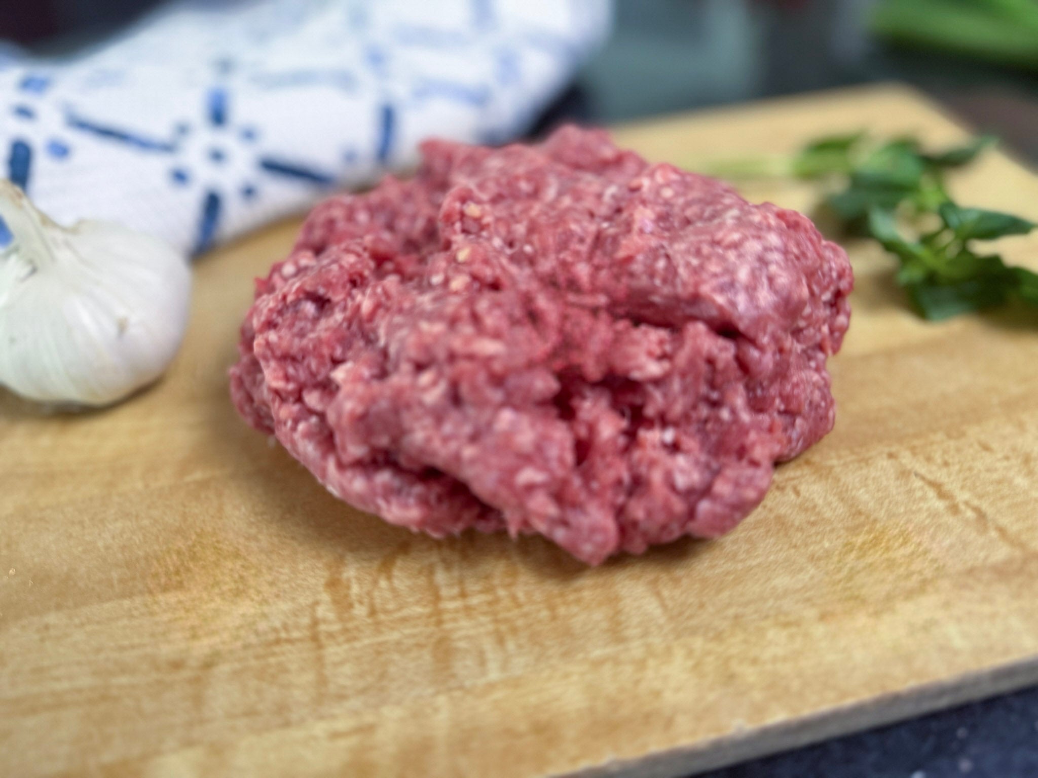 Raw ground meat on a wooden cutting board with garlic and herbs in the background