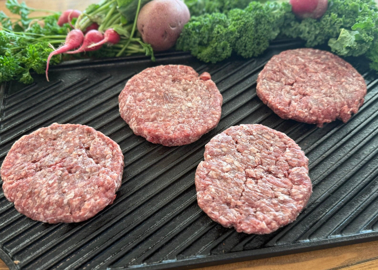 Raw ground beef hamburger patties on a grill pan with fresh vegetables on a wooden surface