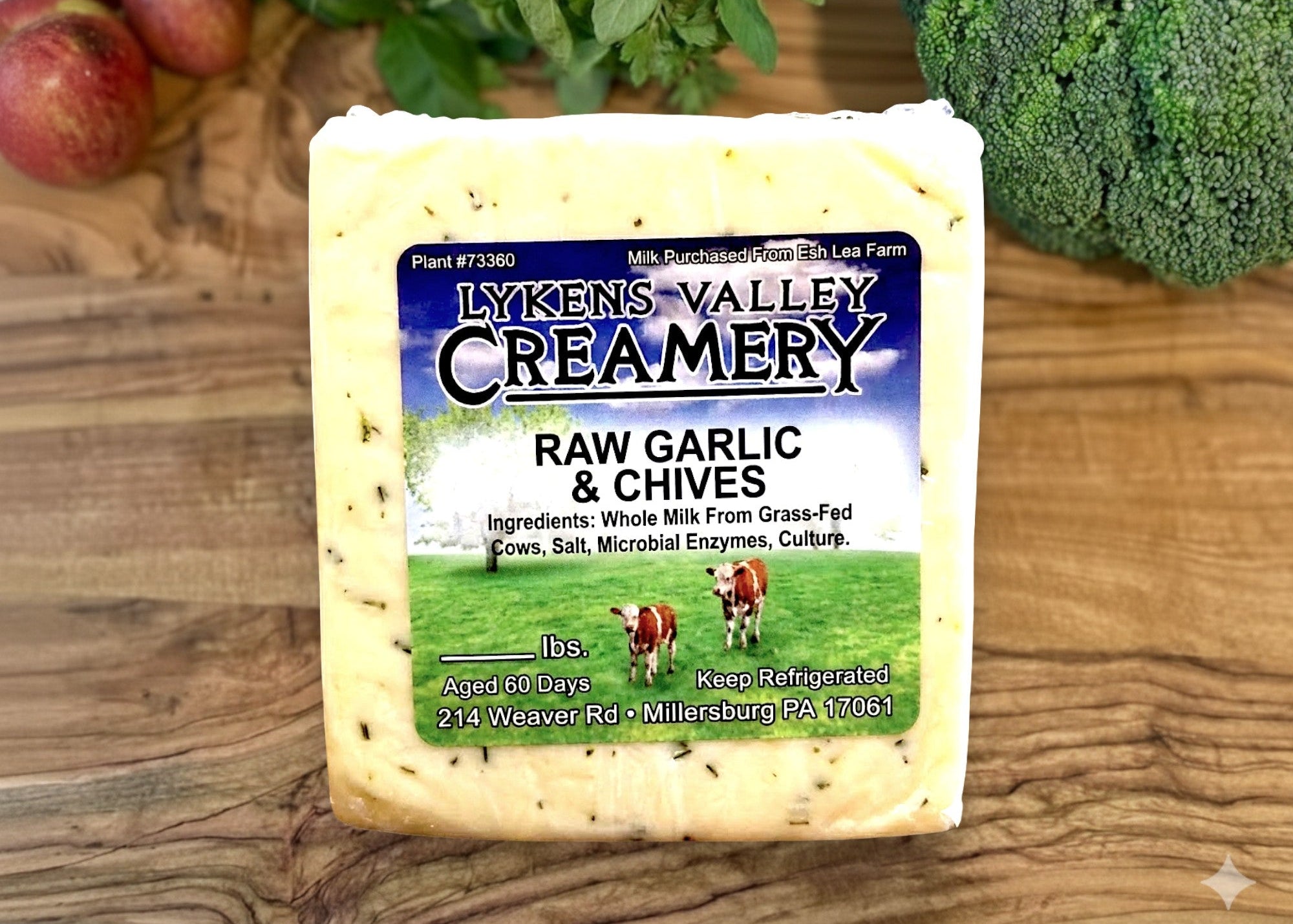 Block of Lyken's Valley Creamery raw garlic & chives cheese on a wooden surface with vegetables in the background.